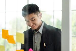 Young adult business asian man write a message and stick on a glass board. photo
