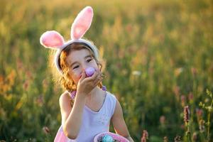 Cute funny girl with painted Easter eggs in spring in nature in a field with golden sunlight and flowers. Easter holiday, Easter bunny with ears, colorful eggs in a basket. Lifestyle photo