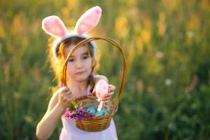 Cute funny girl with painted Easter eggs in spring in nature in a field with golden sunlight and flowers. Easter holiday, Easter bunny with ears, colorful eggs in a basket. Lifestyle photo