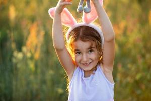 linda niña de 5 años con orejas de conejo abraza suavemente a un conejo de juguete en la naturaleza en un campo floreciente en verano con luz dorada del sol. pascua, conejito de pascua, niñez, niño feliz, primavera. foto