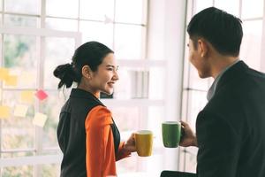 una mujer asiática adulta joven sostiene una taza de café y se relaja hablando con un hombre en la oficina el día. foto