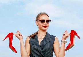 retrato de mujer joven blanca con estilo en traje, gafas de sol, de pie con los brillantes zapatos de tacones altos en sus manos al aire libre foto