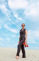 full height portrait of stylish white young woman in suit, sunglasses, red lipstick standing with red glossy high heels shoes in her hands outdoor in desert photo