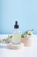 Glass dropper bottle on podium with blooming twigs on blue background. Vertical stock photo