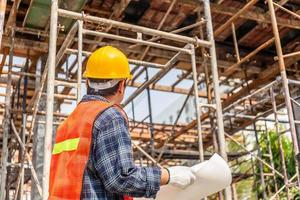 hombre trabajador con sombrero duro que sostiene el proyecto de planificación y verificación de planos en el sitio de construcción foto