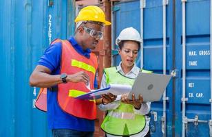 hombre trabajador con casco y chaleco de seguridad sosteniendo una lista de verificación del portapapeles y una capataz usando el control de una computadora portátil cargando la caja de contenedores de la carga foto