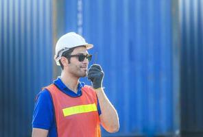 Happy foreman in hardhat and safety vest talks on two-way radio control loading containers box from cargo photo