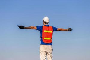 Successful young man with arms wide open, Happy engineer foreman in hardhat and safety vest talks over blue sky background photo