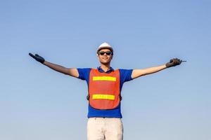 Successful young man with arms wide open, Happy engineer foreman in hardhat and safety vest talks over blue sky background photo