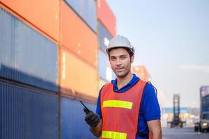 Smiling foreman in hardhat and safety vest talks on two-way radio control loading containers box from cargo photo