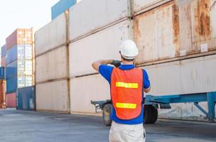 Foreman worker in hard hat and safety vest talks on two-way radio control loading containers box from cargo photo