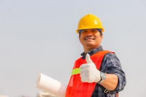 Smiling engineer, worker man holding blueprint with giving thumb up at construction site photo