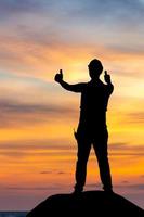 Silhouette of Factory worker man with clipping path in hard hat with thumbs up, young man celebrates with hand in the air sunset background photo