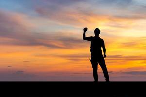 silueta de un trabajador de fábrica con un camino recortado en un sombrero duro con la celebración de la victoria, un joven celebra con la mano en el fondo del atardecer foto