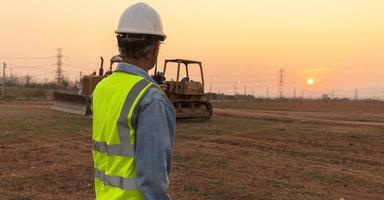 An engineer or worker wearing a life jacket and wearing a white helmet carries a radio transmitter. for safety working on sunset background and crawler bulldozer photo