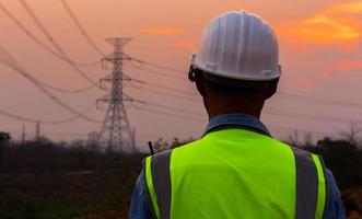 Rear view of an engineer wearing a hard hat in front of a high-voltage electric pole at dusk. photo