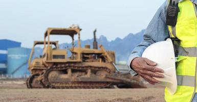 An engineer or worker wearing a safety vest holds a radio transmitter and a white helmet in his hands. for safety working on sunset background and crawler bulldozer photo