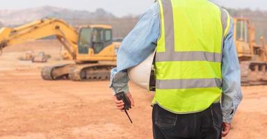 un ingeniero o trabajador que usa un chaleco salvavidas sostiene un transmisor de radio y un casco blanco en sus manos. para un trabajo seguro en el fondo de la excavadora foto