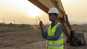 un ingeniero senior con casco y chaleco salvavidas con un transmisor de radio portátil y sobre un fondo de puesta de sol. foto