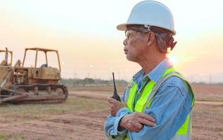 vista lateral, un ingeniero senior masculino asiático que usa chaleco y casco inspecciona las obras civiles, el ingeniero jefe de construcción usa un walkie-talkie para administrar.