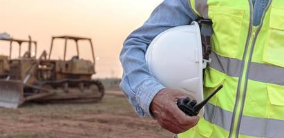 un ingeniero o trabajador que usa un chaleco de seguridad sostiene un transmisor de radio y un casco blanco en sus manos. para la seguridad trabajando en el fondo de la puesta del sol y la excavadora sobre orugas foto