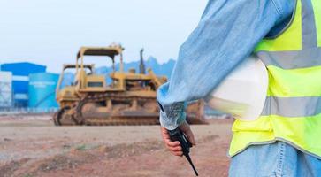 An Asian male engineer or worker wearing a vest holding a radio transmitter and a helmet, a middle-aged foreman controls the excavator. photo