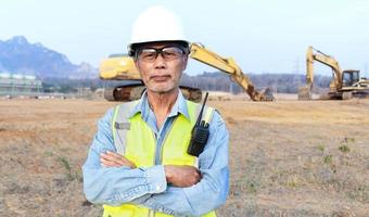 El ingeniero asiático de alto nivel que usa chaleco de seguridad y casco inspecciona las obras civiles. El ingeniero jefe de construcción usa un walkie-talkie para manejar la excavadora.
