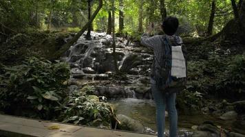 Woman tourist standing on wooden bridge and taking picture with beautiful cascade in tropical rainforest. video