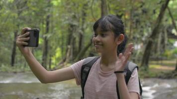 Happy female teenage taking selfie video with smartphone near water stream in tropical forest.