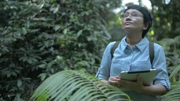 asiatische botanikerin, die an einem digitalen tablet arbeitet, während sie die pflanzenarten für die forschung im wald untersucht.