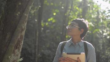 Female biologist looking around in tropical forest and working with digital tablet under morning sunlight. video