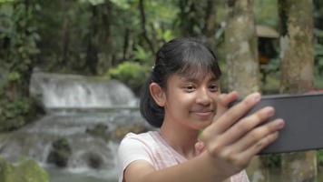 una chica alegre disfruta tomando un video selfie con un smartphone cerca de una hermosa cascada tropical.