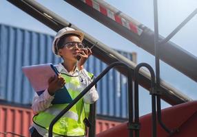 Woman Foreman in hardhat and safety vest holding holding clipboard checklist and talks on two-way radio control loading containers box from cargo photo