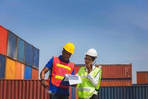 Worker man in hardhat and safety vest holding clipboard checklist and Female foreman talks on two-way radio control loading containers box from cargo photo