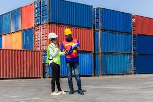 Worker man in hardhat and safety vest holding clipboard checklist and Female foreman talks on two-way radio control loading containers box from cargo photo