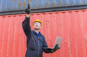 Worker man in hardhat and safety vest holding laptop and point to the sky, Foreman control loading containers box from cargo photo