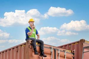 Foreman worker in hardhat and safety vest sitting on container box talks on two-way radio for control loading containers box from cargo photo