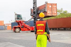 Foreman worker in hardhat and safety vest holding clipboard checklist and two-way radio control loading containers box from cargo photo