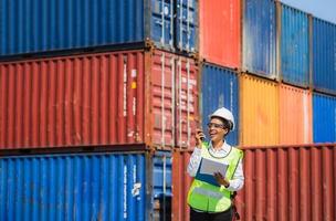 Woman Foreman in hardhat and safety vest holding clipboard checklist and talks on two-way radio control loading containers box from cargo photo