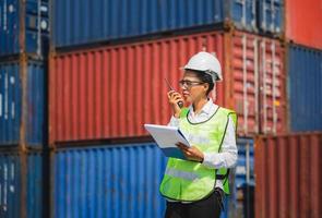 Woman Foreman in hardhat and safety vest holding holding clipboard checklist and talks on two-way radio control loading containers box from cargo photo