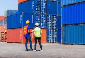 Female foreman with safety vest holding laptop and  worker man in hardhat checking and control loading containers box from cargo photo