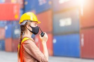 Woman Foreman in hardhat and safety vest holding holding clipboard checklist and talks on two-way radio control loading containers box from cargo photo