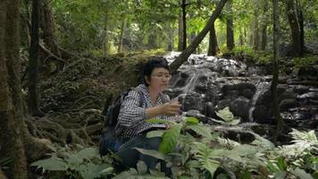 Female botany researcher examining and learning characteristics of plants that grow around waterfall. video