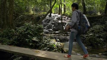 Female tourist walking on wooden bridge through the forest and taking picture with beautiful scenery of cascade.