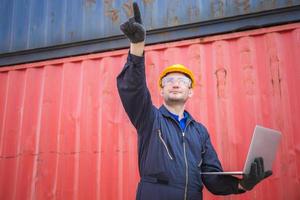 Worker man in hardhat and safety vest holding laptop and point to the sky, Foreman control loading containers box from cargo photo