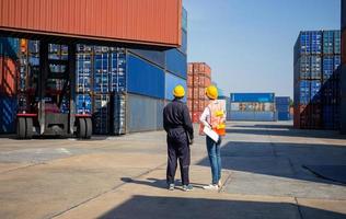 Worker man in hardhat holding laptop and Female foreman with safety vest checking and control loading containers box from cargo photo