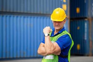 Happiness foreman worker in hardhat show strong arm muscles at containers cargo on a hot day photo