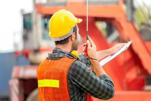 Foreman in hardhat and safety vest holding clipboard checklist and talks on two-way radio control loading containers box from cargo photo