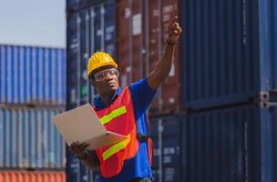 Worker man in hardhat and safety vest holding laptop and point to the sky, Foreman control loading containers box from cargo photo