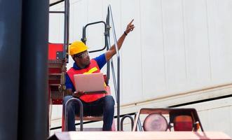 Worker man in hardhat and safety vest holding laptop on container stackers control loading containers box from cargo photo